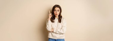 Young woman suggests an idea, raising finger, has revelation, pitching a plan, pointing up, standing over beige background.