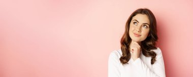 Close up portrait of coquettish smiling woman, glamour girl thinking, looking thoughtful, standing over pink background.
