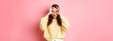 Şaşırmış genç bir kadın nefes nefese kalmış, tanıtım teklifini kontrol ediyor, el dürbününden bakıyor, pembe arka planda duruyor..