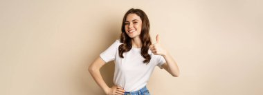 Modern genç bir kadının portresi başparmaklarını kaldırıyor, onaylıyor, gülümsüyor, memnun oluyor, şirket ya da web sitesi tavsiye ediyor, bej arka planda duruyor..