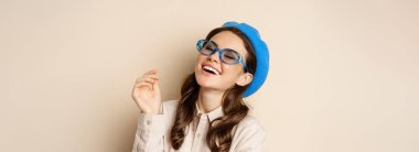 Close up portrait of stylish woman posing in sunglasses, laughing and smiling, wearing trendy outfit, standing over beige background.