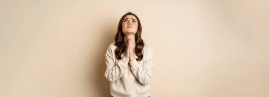 Woman begging and pleading god, supplicating, say please, standing desperate over beige background. Copy space