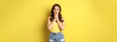 Shocked young woman gasping, looking amazed at camera, standing in tank top over yellow background. Copy space