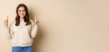 Enthusiastic girl pointing fingers up, showing announcement, promo text or logo upwards, standing over beige background.
