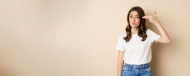 Young woman showing finger pistol sign near head and looking annoyed, sick and tired, standing over beige background.