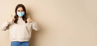 Covid-19, salgın ve karantina konsepti. Genç kadın Coronavirus omicron salgını sırasında tıbbi yüz maskesi takıyor, baş parmaklarını gösteriyor, bej arka planda duruyor..