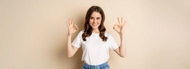 Beautiful female model smiling pleased, showing okay, zero sign, complimenting, praising you, standing over beige background.