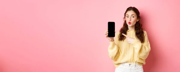 stock image Technology and online shopping. Excited girl looks curious, points finger at empty mobile phone screen, shows app on smartphone, standing against pink background.