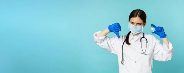 stock image Sad and disappointed young doctor, woman showing vaccine and dislike, thumbs down gesture, disapprove medical drug, standing over blue background.