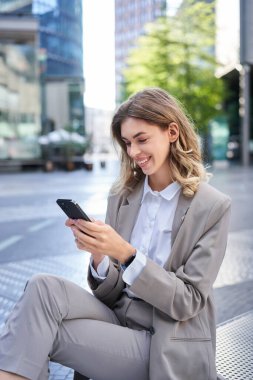 Akıllı telefon uygulamasına bakıp gülümseyen bir iş kadınının dikey görüntüsü, cep telefonuyla mesaj göndermesi, ofis binasının dışında otururken yemek yemesi..