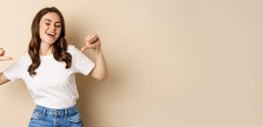 Self-assured young happy woman pointing fingers at herself and dancing, self-promoting, being confident, standing over beige background.