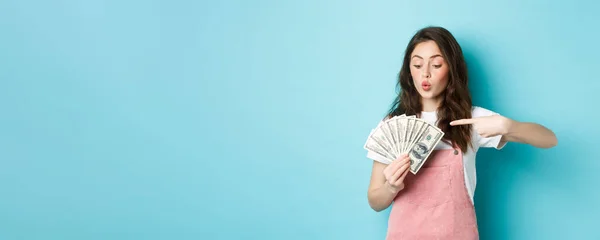 stock image Excited cute girl holding money and pointing at dollar bills, going on shopping, standing over blue background.