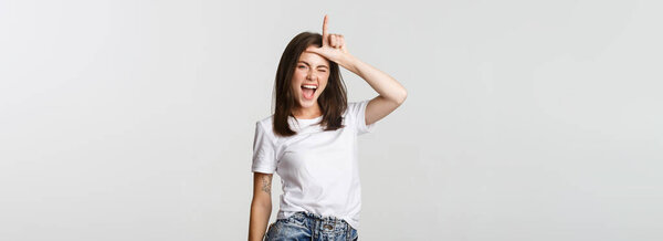 Sassy good-looking girl smiling and mocking person, showing loser gesture on forehead.