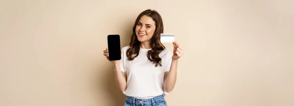 オンラインショッピングや人々の概念 若い美しい女性が幸せそうに見えます クレジットカードの割引や携帯電話の画面を表示 ベージュの背景の上に立って — ストック写真