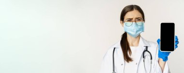 Online medical help concept. Sad and gloomy young doctor in face mask, showing smartphone screen with upset an disappointed expression, white background.