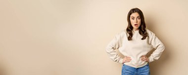 Portrait of shocked brunette woman drop jaw, gasping and staring speechless at camera, beige background. Copy space