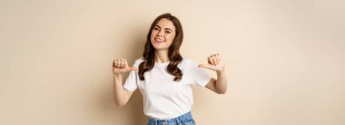 Self-assured young happy woman pointing fingers at herself and dancing, self-promoting, being confident, standing over beige background.