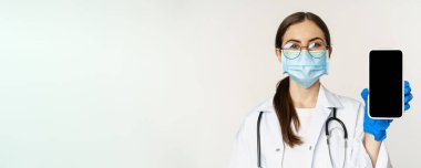 Online medical help concept. Woman doctor in glasses and face mask, showing mobile phone screen, app interface or website for patients, standing over white background.