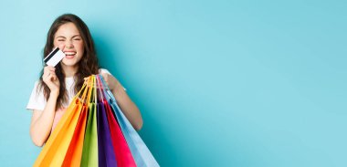 Mutlu genç bir kadın gülüyor, alışveriş torbaları ve plastik kredi kartları gösteriyor, indirimli mağazalardan alışveriş yapıyor, mavi arka plana karşı neşeli duruyor..