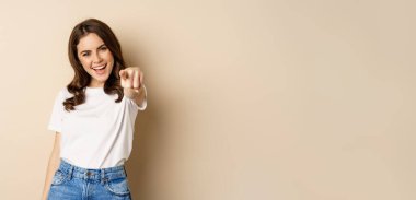 Its you, congrats. Smiling beautiful woman pointing finger at camera, congratulating, praising you, standing over beige background.