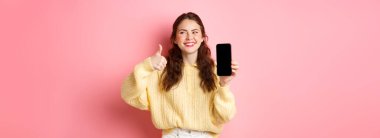 Güzel bir kadının gülüşü ve yan bakışları, bir uygulamayı derecelendirirken, başparmaklarını kaldırıp boş bir telefon ekranı göstermesi, online tanıtım önermesi, pembe arka plana karşı durması.