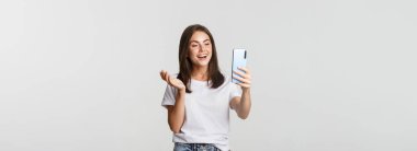 Arkadaş canlısı, çekici bir kız arkadaş, gülümseyen ve sohbet eden, akıllı telefon tutan, beyaz arka plan.