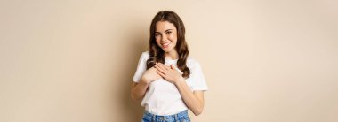 Portrait of young caucasian woman hold hands on heart and smiling, daydreaming, thinking of smth heartwarming, posing against beige background.