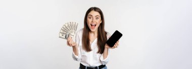 Enthusiastic young woman winning money, showing smartphone app interface and cash, microcredit, prize concept, standing over white background.