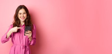 İnternette reklam yapan beyaz bir kız akıllı telefonuna işaret ediyor ve gülümsüyor. Tatmin olmuş, uygulama öneriyor, pembe arka planda duruyor..