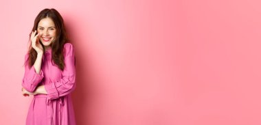 Güzellik ve moda. Çekici beyaz kadın kameraya kıkırdıyor ve gülümsüyor, güzel bir bahar gününde romantik parlak elbiseler içinde pembe arka planda duruyor..