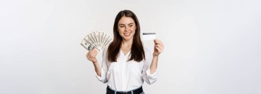 Happy modern woman holding money and credit card in hands, smiling pleased, standing over white background. Copy space