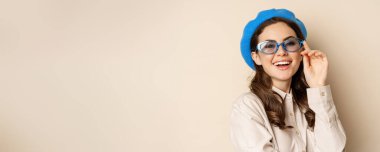 Close up portrait of stylish woman posing in sunglasses, laughing and smiling, wearing trendy outfit, standing over beige background.