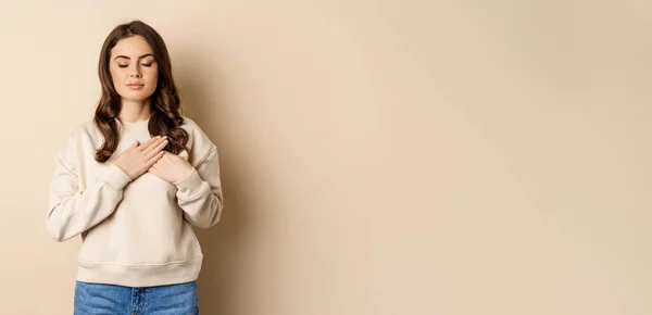 stock image Caring woman close eyes, hold hands on heart, thinking of someone, daydreaming, standing over beige background.