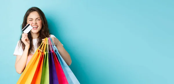 快乐的年轻女人笑着 展示购物袋和塑料信用卡 在打折商店里买东西 站在蓝色的背景下兴高采烈 — 图库照片