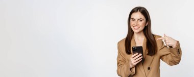Enthusiastic saleswoman, business woman pointing finger at mobile phone and smiling, showing on cellphone, standing against white background.