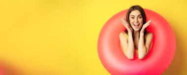 Happy beautiful young woman relaxing on summer vacation, wearing pink swim ring for beach holiday, standing against yellow background.