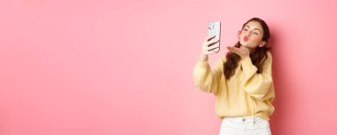 Güzel flörtçü kız akıllı telefondan selfie çekiyor, görüntülü konuşma sırasında hava öpücüğü gönderiyor, pembe arka planda duruyor. Boşluğu kopyala