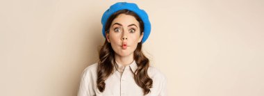 Young funny woman acting, fooling around, pucker lips and grimacing, posing against beige background in trendy blue hat.