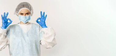 Image of healthcare worker, doctor in personal protective equipment from covid-19, showing okay ok sign, confident face in mask, standing over white background.