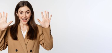 Enthusiastic businesswoman in suit, raising hands palms up, number ten dozen gesture and smiling, standing over white background.