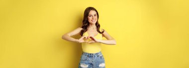 Beautiful young woman showing heart, love sign, like gesture, smiling happy and cute, standing in tank top and shorts over yellow background.