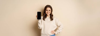 Stylish smiling woman showing smartphone screen, mobile app interface, standing over beige background.