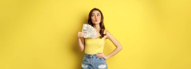 Shopping, credit and money concept. Young brunette woman showing cash and smiling pleased, standing over yellow background.