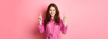 Tarz sahibi, kıvırcık saçlı, açık renk elbisesi olan, gülümseyen, logoyu işaret eden, fotokopi alanını gösteren, pembe arka planda duran beyaz bir kadın..