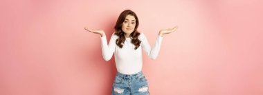 I dont know. Confused cute young woman smiling awkward, shrugging shoulders clueless, has no idea, standing over pink background.