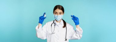 Gülümseyen sağlık personeli, maske takan doktor ve lastik eldiven, mavi arka planda duran covid-19 salgınından şırınga ve aşı gösteriyor..