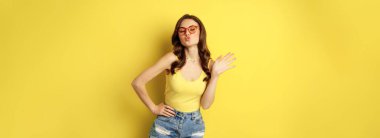 Stylish pretty woman posing in sunglasses and summer clothes, looking happy and carefree, concept of vacation and sunny days, standing over yellow background.