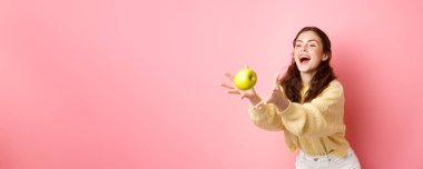 Mutlu tatlı kız bir elma yakalar ve güler, sağlıklı öğle yemeği zamanı meyvelerle dolu, pembe arka planda durur.