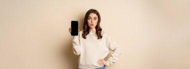 Stylish smiling woman showing smartphone screen, mobile app interface, standing over beige background.