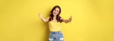 Beautiful girl in tank top, reaching hands, stretching arms forward and smiling, welcome, hugging or greeting someone, standing over yellow background.